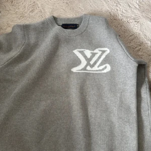 Grå stickad tröja Louis Vuitton - Snygg grå stickad tröja från Louis Vuitton med stort vitt LV-logo på bröstet. Rund hals och långa ärmar ger en clean och modern look. Perfekt för dig som gillar exklusiva märken och vill ha något som sticker ut i garderoben. Storlek M