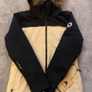 Burton beige och svart parkajacka - Snygg parkajacka från Burton i beige och svart med huva och fuskpäls. Jackan har dragkedja framtill, praktiska fickor och logga på ärmen. Perfekt för kalla dagar och har en clean, sportig look som funkar till streetwear.