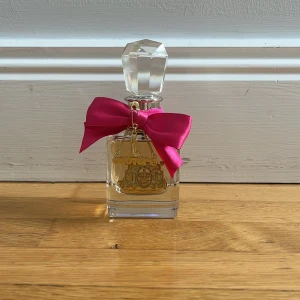 Juicy Couture parfym med rosa rosett - Juicy Couture parfym i en lyxig, fyrkantig glasflaska med guldfärgad logga och detaljer. Flaskan har en stor, kristalliknande kork och dekoreras med en rosa satinrosett och guldfärgad 'Couture'-berlock. Perfekt för dig som gillar glam och statement-detaljer.