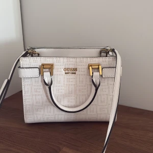 Vit handväska från Guess med guldiga detaljer - Snygg vit handväska från Guess med präglat mönster och guldiga metalldetaljer. Väskan har både handtag och axelrem, samt flera fack och dragkedja inuti. Perfekt storlek.