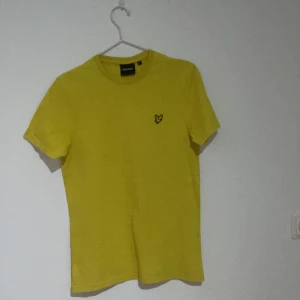Gul t-shirt från Lyle & Scott - Snygg gul t-shirt från Lyle & Scott med klassisk rund hals och korta ärmar. På bröstet sitter den ikoniska fågel-loggan i svart och gult. T-shirten är gjord i mjuk bomull och har en clean, stilren look som passar perfekt till jeans eller shorts.