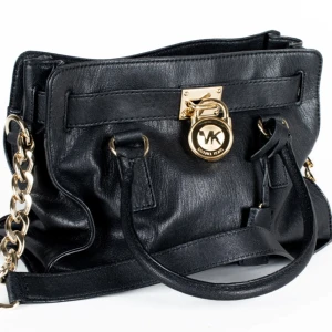 Svart handväska från Michael Kors - Snygg svart handväska från Michael Kors i skinn med guldfärgade kedjedetaljer och lås framtill. Väskan har två handtag, axelrem och ett rymligt innerfack med dragkedja. Insidan är beige med MK-logga. Perfekt för dig som gillar lyxiga detaljer. Äkta köpt i Dubai. Välvårdad och inga fläckar i fodret. Hämtas helst i Stockholm Solna alt. Kan skicka mot Swish i samband med att jag postar. Köparen betalar frakt