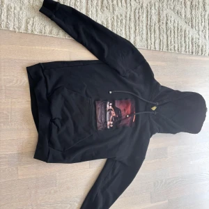 Svart hoodie med tryck och huva - Svart hoodie med stor ficka framtill och coolt tryck av en person på en bil med röd bakgrund. Tröjan har huva med snörning och långa ärmar. Perfekt för dig som gillar streetstyle och vill sticka ut med ett unikt motiv.