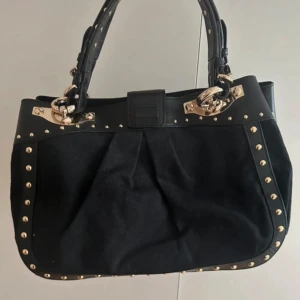 Svart väska med gulddetaljer från DKNY - Snygg svart väska från DKNY med guldfärgade nitar och detaljer. Väskan har dubbla handtag i skinn och ett diskret mönster i tyget. Perfekt storlek för att få plats med allt du behöver