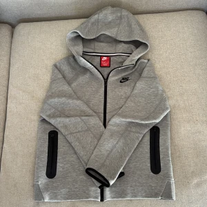 Grå hoodiejacka från Nike S - Snygg grå hoodiejacka från Nike i storlek s med svart logga på bröstet och hel dragkedja framtill. Jackan har två svarta zip-fickor på sidorna och en mjuk, sportig känsla. Perfekt för chill eller träning, med skön huva och stilren look. Passar dig runt 1,70. Finns ett lite hål på ryggen men de syns inte och går att laga.