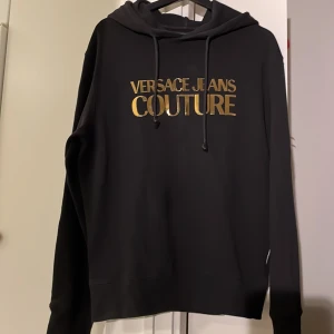 Svart hoodie Versace Jeans Couture - Svart hoodie från Versace Jeans Couture med stor, glansig guldfärgad logga på bröstet. Tröjan har huva med snörning. Vissa bokstäver är lösa men kan fästas med textillim. Använd 2-3 gånger. 