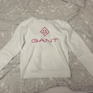 Gant hoodie - Vit hoodie från GANT med rosa logga och tryck på bröstet. Tröjan har huva med snörning, magficka och långa ärmar. Mjuk och skön bomullskvalitet, perfekt för chill dagar. Snyggt kontrasttryck i rosa som ger en fräsch vibe. Jätteskönt material, speciellt inuti. Tröjan har en fläck på fickan, men det syns knappt och tror det går att få bort den om man försöker. Men pågrund av fläcken så är priset lägre.
