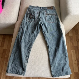 Blå cargopants från Blue Wear med tryck - Säljer ett par blå cargopants från Blue Wear med coola detaljer som broderad fågel på bakfickan och stora svarta patchar med döskalle- och texttryck på framsidan. Byxorna har röda sömmar, dragkedja på bakfickan och flera fickor. Perfekt för dig som gillar streetstyle. 