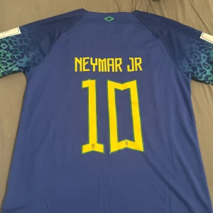 Blå Brasil Neymar Jr 10 Nike tröja - Säljer en officiell Brasilien fotbollströja från Nike med Neymar Jr och nummer 10 på ryggen. Tröjan är blå med gula detaljer och har korta ärmar med leopardmönster i mörkblått och grönt. Materialet är lätt och andas, perfekt för match eller träning. 