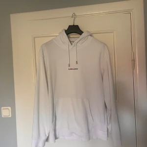 Vit hoodie från Saint Laurent - Säljer en clean vit hoodie från Saint Laurent med svart och röd logga på bröstet. Tröjan har huva med snörning, långa ärmar och en stor magficka. Materialet är mjukt och känns som bomull, perfekt för chill / kyligare dagar. Metalländarna på snörena har Saint Laurent-gravyr. Sitter mer som en M , hör bara av dig om några frågor eller funderingar!!