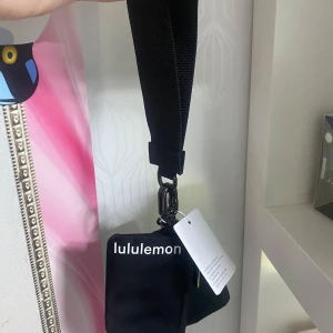 Hell ny Svart axelväska från Lululemon - Säljer en hell ny, cool svart axelväska från Lululemon med bred justerbar rem och två separata fack med dragkedja. Väskan har stilren design med vit logga framtill och metallspännen. Perfekt för att bära dina essentials och hålla stilen. En hell ny från nätet kostar 480 kr, mitt pris 350 kr.