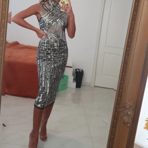 Glittrig silverklänning med paljetter - Säljer en superglittrig silverfärgad klänning med paljetter och coola detaljer. Klänningen är ärmlös, har hög hals och ett korslagt mönster över bröstet. Den är figurnära och går ner till vaden, perfekt för att sticka ut på festen. Köpt 1800kr 