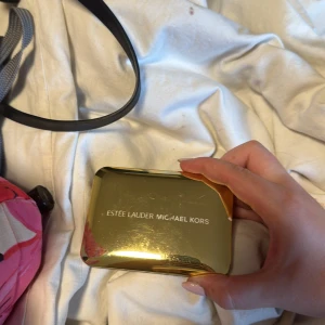 Estée Lauder Michael Kors sminkpalett - Snygg sminkpalett i guldig förpackning med spegel. Innehåller sex ögonskuggor i lila, brunt, beige och skimrande nyanser samt en matt bronzer. Perfekt för att skapa både mjuka och dramatiska looks. Paletten är kompakt och enkel att ta med i väskan.