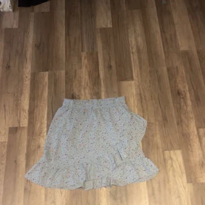 Blommig volangkjol från Pieces - Supersöt ljusblå kortkjol från Pieces med blommigt mönster i lila, rosa och gult. Kjolen har volangkant nedtill och resår i midjan för skön passform. Perfekt för vår och sommar när du vill ha en chill och feminin look.