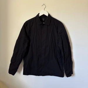 Svart overshirt från COS, strl 50 - Svart overshirt från COS i storlek 50. Skjortan har klassisk krage, raka linjer och långa ärmar. Materialet är en mix av bomull och polyamid, vilket gör den lätt och bekväm. Perfekt för en clean och stilren look. 1C11