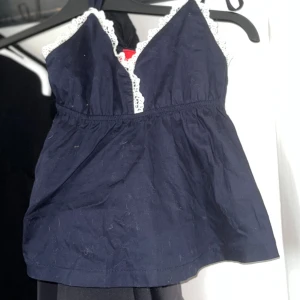 Mörkblå linnetopp med spets från RYVLS - Supersöt mörkblå linnetopp från RYVLS med vita spetsdetaljer längs v-ringningen och axelband som knyts. Toppen har en lätt rynkad midja och ärmlös design, perfekt för att ge en chill och feminin vibe. Materialet känns mjukt och luftigt, idealiskt för varmare dagar.