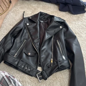 Svart skinnjacka med bälte från Zara  - Snygg svart skinnjacka från Zara  med klassisk bikerstil. Jackan har silvriga dragkedjor, knappar och ett brett bälte med metallspänne i midjan. Flera fickor med dragkedja och detaljer som ger en cool vibe. Perfekt för dig som vill ha en edgy look.