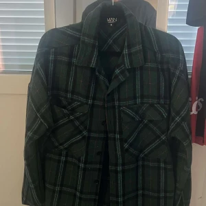 Grön rutig overshirt från MAN - Snygg grön overshirt från MAN i storlek M med rutigt mönster i mörkgrönt, svart och tunna vita linjer. Skjortan har långa ärmar, klassisk krage, två bröstfickor med knappar och knäppning framtill. Perfekt för lager-på-lager och chill stil.