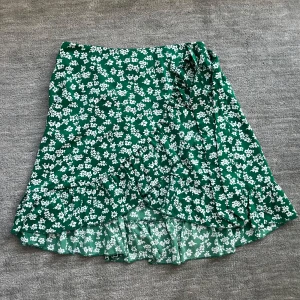 Grön volangkjol med vitt blommönster  - Säljer nu denna gröna volangkjol med vitt blommönster på. Kjolen har två band som man kan knyta till en rosett. Kjolen köptes för 120 kr men säljer den nu för 49 kr. Endast använd 1 gång!