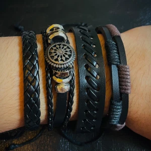 Svart läderarmband med detaljer - Snyggt set med flera svarta armband i läder och textil, vissa flätade och andra med coola snörningsdetaljer. Ett armband har en rund metallbricka med mönster och silverfärgade pärlor. Perfekt för dig som gillar edgy och unik stil.