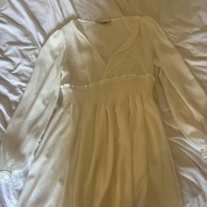 Vit omlottklänning med smock - Supersöt vit kortklänning med omlott upptill och smockad midja som ger en snygg siluett. Klänningen har långa, lätt transparenta ärmar med resår vid ärmslut och volangdetalj vid midjan. Perfekt för sommarens alla tillfällen!