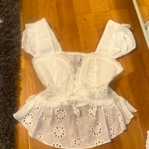 Vit blus med broderade detaljer - Superfin vit blus från Amisu med broderade blommor och volangkant. Toppen har puffiga korta ärmar, omlott framtill med knyt och en söt v-ringning. Materialet är lätt och luftigt bomull, perfekt för varma dagar.