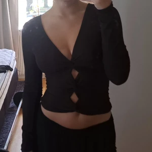 Svart ribbad omlottop - Säljer en svart långärmad topp med ribbad struktur och djup v-ringning. Toppen har tre snygga knyt framtill som skapar öppna partier och ger en cool omlott-effekt. Passformen är tight och toppen är croppad, perfekt för att lyfta din outfit.