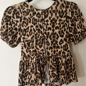 Leopardmönstrad blus från Gina Tricot - Säljer en beige och svart blus från Gina Tricot med leopardmönster. Toppen har puffiga korta ärmar, volang nedtill och snygga knytningar där fram. Perfekt för dig som vill sticka ut med ett djuriskt print och coola detaljer.