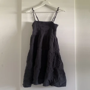 Svart klänning med smockliv - Svart klänning med smala justerbara axelband och smockat liv. Klänningen har en luftig, vågig kjol och är tillverkad i ett mjukt, strukturerat bomullstyg. Perfekt för dig som gillar en enkel men stilren look.