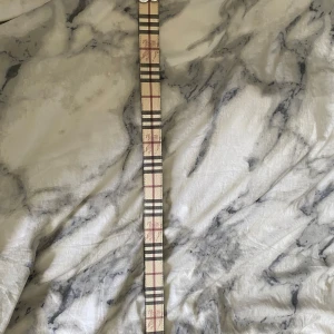 Rutig Burberry bälte i skinn - Snyggt Burberry bälte i skinn med klassiskt rutigt mönster i beige, svart och vinröd. Bältet har en silverfärgad metallspänne med Burberrys B-logga och präglad riddare på båda sidor. Perfekt accessoar för att lyfta din outfit.