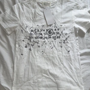 Vit t-shirt Maison Margiela med print - Snygg vit t-shirt från Maison Margiela med ikoniskt sifferprint och gråa färgstänk framtill. Klassisk passform, rund hals och korta ärmar. Tillverkad i mjuk bomull som känns skön mot huden. Perfekt för dig som gillar streetstyle med en twist.