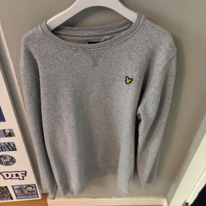 Grå sweatshirt från Lyle & Scott - Snygg grå sweatshirt från Lyle & Scott med klassisk rund hals och den ikoniska gula loggan broderad på bröstet. Tröjan har långa ärmar och ribbade muddar vid ärmslut och nederkant. Perfekt för en chill och stilren look.