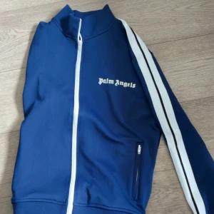 Blå Palm Angels traksuits kofta  S - Säljer en blå zip ett set ifrån traksuiten från Palm Angels med vita ränder längs ärmarna och vit dragkedja. Jackan har hög krage, fickor med dragkedja och logga på bröstet. Tillverkad i polyester och har en sportig vibe. Perfekt för dig som gillar streetwear.