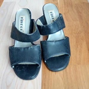 Svarta sandaler med klack från Firence - Svarta sandaler från Firence med breda remmar och öppen tå. Skorna har en stabil hög klack och platt sula framtill. Tillverkade i syntetmaterial och passar perfekt till varmare dagar när du vill ha en stilren look.Storleken är 37