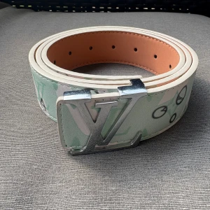 Louis Vuitton bälte med silverfärgat spänne - Snyggt Louis Vuitton bälte med bred design och ikoniskt LV-spänne i silver. Bältet har ett ljust mönster i vitt och mintgrönt med klassiska LV-loggor och detaljer. Tillverkat i skinn med ljus insida och exklusiv känsla. Perfekt för att lyfta din outfit.