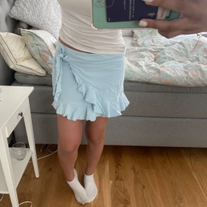 Ljusblå omlottkjol med volang - Supersöt ljusblå kortkjol med omlottmodell och volangkant. Kjolen har en knytning i sidan som ger en extra detalj och är gjord i ett mjukt bomullstyg. Perfekt för dig som gillar en feminin och chill stil.