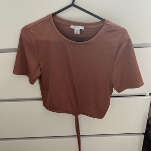 Ribbad brun croppad topp - Säljer en ribbad brun croppad topp i storlek S. Toppen har korta ärmar och en snygg knyt-detalj framtill som ger en trendig look. Perfekt för dig som gillar en enkel men cool stil.