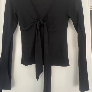 Svart långärmad knytblus Gina Tricot - Supersnygg svart långärmad topp från Gina Tricot med v-ringning och knyt framtill. Toppens material är mjukt och stretchigt, perfekt för en trendig look. Passformen är slim och ärmarna är långa för en stilren vibe. Har används en gång eftersom jag köpte fel storlek, skriv om du är intresserad eller om mer info