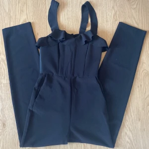 Svart byxdress med volangdetalj från H&M storlek 34 - Snygg svart byxdress med breda axelband och volang upptill. Byxdressen har raka ben och dragkedja i ryggen för enkel påklädning. Strukturerad vid bysten och har 2 fickor. Tillverkad i ett mjukt, vävt material som ger en stilren look. Perfekt för dig som vill sticka ut med en trendig och bekväm outfit.