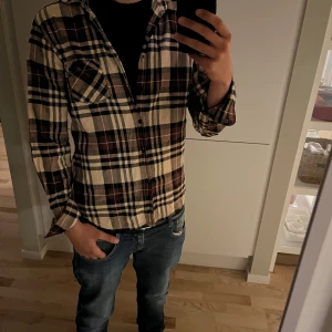 Rutig skjorta från Replay. - Säljer en snygg rutig skjorta från Replay i regular fit. Skjortan har bruna, beige och svarta rutor, klassisk krage och knäppning framtill. Långärmad modell med bröstficka, perfekt att bära öppen över en t-shirt eller stängd.