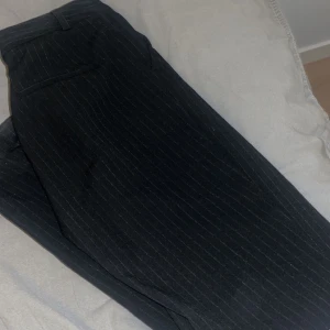 Svarta kostymbyxor med smala ränder - Snygga svarta kostymbyxor med tunna vita ränder. Klassisk passform med fickor bak och bälteshällor. Perfekta för en stilren look och passar till skjorta eller t-shirt.