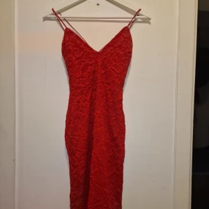 Röd spetsklänning med smala axelband - Snygg röd klänning med smala axelband och hjärtformad uringning. Klänningen är täckt av vacker spets med blommigt mönster och har en figurnära passform. Perfekt för dig som vill sticka ut med en färgstark look.