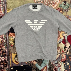 Grå sweatshirt från Emporio Armani - Säljer en stilren grå sweatshirt från Emporio Armani med det klassiska vita logotrycket framtill. Tröjan har rund halsringning och långa ärmar. Perfekt för dig som gillar en clean och exklusiv look.