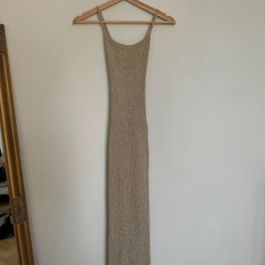 Glittrig guld långklänning med snörning - Lngklänning i glittrigt guldigt material med smala axelband och figurnära passform. Klänningen har snörning i ryggen som ger en cool detalj och gör att den sitter perfekt. 