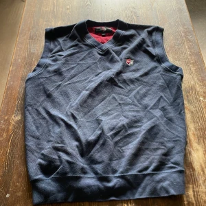 Mörkblå väst från Wilson Staff - Mörkblå väst från Wilson Staff med v-ringning och liten logga på bröstet. Västen är i bomull och har en klassisk, sportig look med ribbade kanter. Perfekt för lager-på-lager och passar till både jeans och chinos.