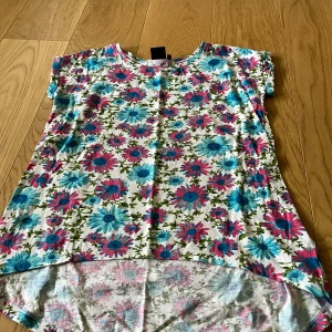 Blommig topp från N.T.Y. i bomull - Superfin topp från N.T.Y. med färgglatt blommönster i blått och rosa på vit botten. Modellen är lite längre bak och har korta ärmar. Perfekt för dig som gillar en lekfull och somrig stil. Mjuk och skön i bomull.