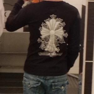 Svart långärmad tröja Chrome Hearts - Svart långärmad tröja från Chrome Hearts med stora vita korsdetaljer på ryggen, ärmen och framtill. Tröjan har en normal passform och är tillverkad i mjuk bomull. Snygg streetwear vibe med tydliga loggor och grafiska tryck.