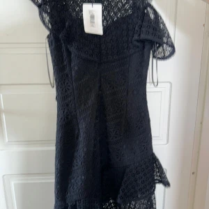 Svart spetsklänning med volanger - Supersnygg svart långklänning i spets med blommigt mönster och volangdetaljer. Klänningen har korta ärmar och transparenta partier upptill för en cool look. Perfekt för dig som vill sticka ut med en elegant och trendig stil. Köpte i USA för 2500kr och säljer för 800