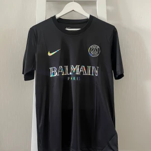 Svart PSG x Balmain x Nike fotbollströja - Svart fotbollströja från Nike i samarbete med PSG och Balmain. Tröjan har holografiska detaljer på Nike-loggan, PSG-emblemet och Balmain Paris-trycket på bröstet. Tillverkad i ett lätt och sportigt material med rund hals och korta ärmar.