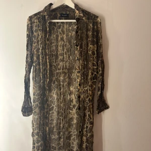 Ormmönstrad kofta från Sand Copenhagen - Snygg långärmad kofta från Sand Copenhagen med ormmönster i bruna och beige toner. Skjortan har knappar framtill och är tillverkad i tunn, luftig viskos. Perfekt för dig som vill sticka ut med ett djurmönstrat plagg. Tunt material som är lite genomskinligt o något liten i storlek